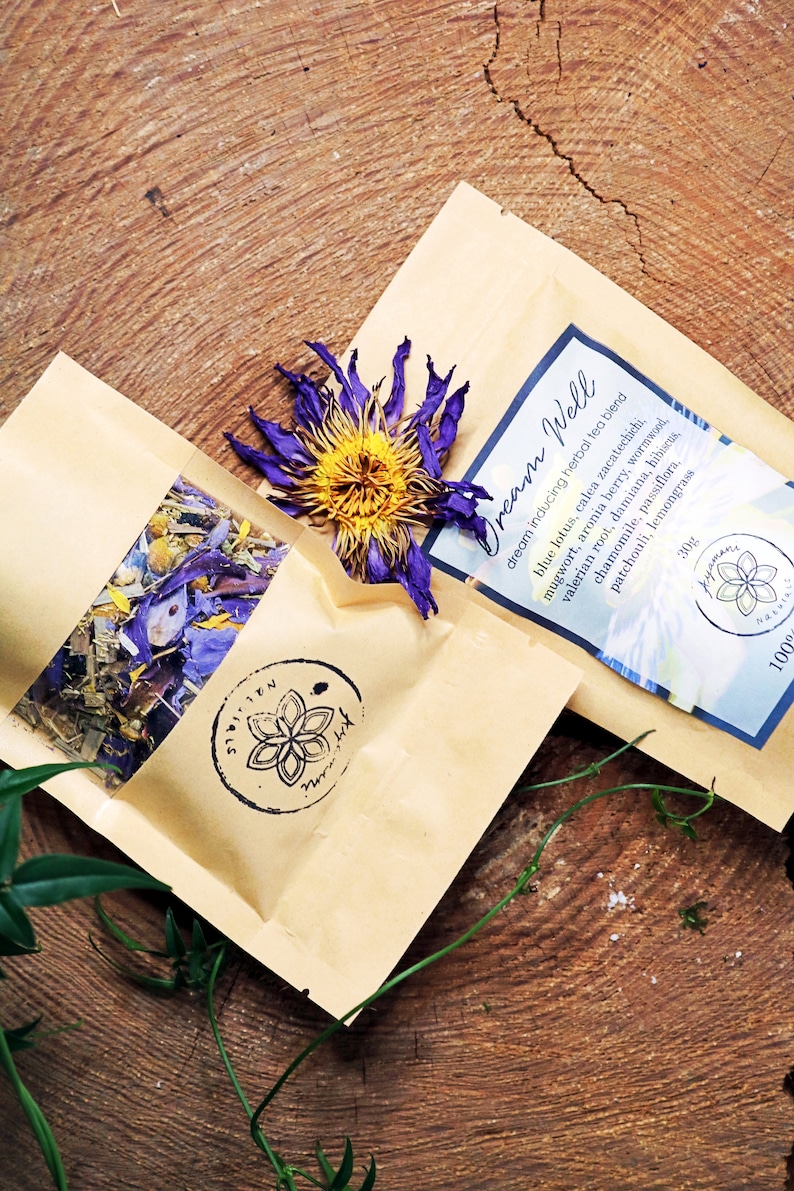 May include: Two brown paper bags with a blue and yellow flower on top. The bag on the right has the text "Dream Well" and lists the ingredients of a herbal tea blend. The bag on the left has a clear window showing a variety of dried herbs.