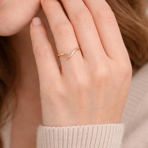May include: A delicate gold-colored ring with a wave design. The ring is worn on a finger, showcasing its simple yet elegant form. The ring's smooth, polished surface reflects light, highlighting its subtle curves and minimalist aesthetic.