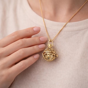 Puede incluir: Un collar con colgante dorado con una esfera de filigrana con intrincados calados. La esfera tiene una pequeña gema visible. El collar está en una delicada cadena dorada.