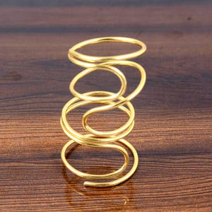 May include: A gold-colored, spiral-shaped ring. The ring is made of a thin, continuous band of metal that coils around itself. The design is simple and elegant, with a polished finish. The ring is shown against a dark brown wooden surface.
