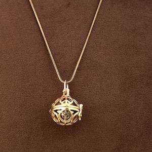Puede incluir: Un collar de cadena dorada con un colgante redondo decorativo en forma de jaula. El colgante tiene un diseño delicado e intrincado y parece ser hueco.