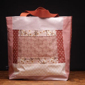 Könnte beinhalten: Eine Patchwork-Tragetasche in Rosa- und Rosttönen. Die Tasche weist verschiedene Muster auf, darunter Punkte, Blumendrucke und geometrische Designs. Sie hat einen rostfarbenen Griff und eine rechteckige Form, die sich zum Tragen von Gegenständen eignet.