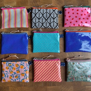 Könnte beinhalten: Neun kleine Reißverschlusstaschen mit verschiedenen Mustern, darunter Streifen, Punkte und florale Designs. Die Taschen haben eine Vielzahl von Farben, wie Rosa, Blau und Grün. Jede Tasche hat eine kleine Schlaufe und einen Reißverschluss.