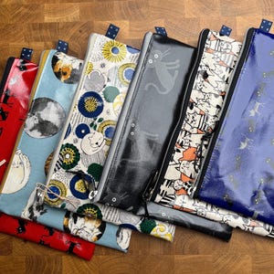 Könnte beinhalten: Sechs rechteckige Reißverschlusstaschen in einer Reihe, jede mit unterschiedlichen Mustern zum Thema Katzen. Die Designs umfassen Rot, Hellblau, Weiß mit Blumenkreisen, Schwarz, Creme mit orangefarbenen Katzen und ein glänzendes Dunkelblau mit Katzensilhouetten.