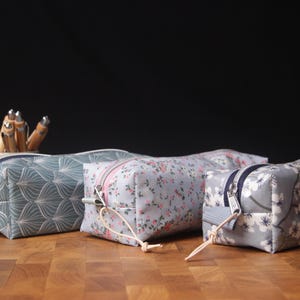 Könnte beinhalten: Drei Federmäppchen aus Stoff mit Reißverschlüssen liegen auf einer Holzoberfläche. Die Etuis weisen verschiedene Muster auf: ein türkisfarbenes und weißes geometrisches Design, ein Blumenmuster und ein graues Blumenmuster. Stifte sind im Hintergrund zu sehen.