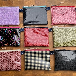 Könnte beinhalten: Neun rechteckige Reißverschlusstaschen in einem Raster angeordnet. Die Taschen zeigen verschiedene Muster, darunter violette, rote und schwarze geometrische Drucke, ein schwarzes Blumenmuster, ein rot-weißes Wellenmuster, ein grünes Wellenmuster und ein schwarzes Vichy-Karo.