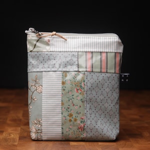 Könnte beinhalten: Eine kleine, gepatchte Reißverschlusstasche mit verschiedenen floralen und geometrischen Mustern in Grün-, Rosa- und Weißtönen. Die Tasche hat einen Reißverschluss und einen Lederzug.