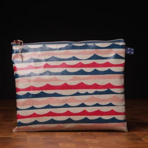Könnte beinhalten: Eine rechteckige Tasche mit einem sich wiederholenden Wellenmuster in Rot, Blau und Rosa. Die Tasche hat einen Reißverschluss und ein kleines Stoffetikett an der Seite. Der Hintergrund ist dunkel und die Tasche liegt auf einer Holzoberfläche.