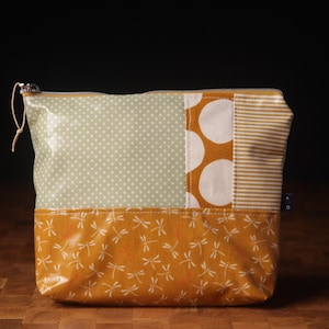 Könnte beinhalten: Ein Patchwork-Reißverschlusstasche mit einer Vielzahl von Mustern. Die obere Hälfte zeigt ein hellgrünes Polka-Dot-Muster und einen Abschnitt weißer Kreise auf senffarbenem Hintergrund. Die untere Hälfte hat ein senfgelbes Libellenmuster.