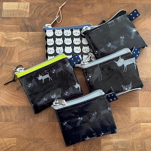 Könnte beinhalten: Eine Sammlung kleiner Reißverschlusstaschen mit Katzenmotiven. Die Taschen haben schwarze Hintergründe mit weißen oder grauen Katzenillustrationen, und eine Tasche hat ein weißes Katzenmuster. Die Reißverschlüsse haben verschiedene farbige Verzierungen.