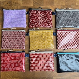 Könnte beinhalten: Neun kleine Reißverschlusstaschen mit Muster in verschiedenen Farben, darunter Lila, Rot, Gelb und Schwarz. Jede Tasche hat ein einzigartiges geometrisches oder wellenartiges Design. Die Taschen sind auf einer Holzoberfläche angeordnet.