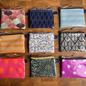 Könnte beinhalten: Neun kleine, quadratische Reißverschlusstaschen mit verschiedenen Mustern und Farben sind auf einer Holzoberfläche angeordnet. Die Taschen weisen Designs wie Blumen-, geometrische und abstrakte Muster in Rosa, Blau, Orange und Schwarz auf.