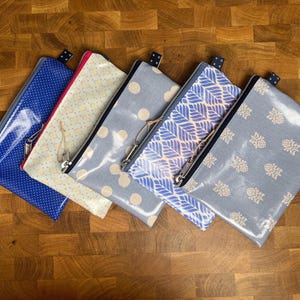 Könnte beinhalten: Fünf Reißverschlusstaschen in verschiedenen Mustern und Farben liegen auf einer Holzoberfläche. Die Taschen umfassen Blau mit weißen Punkten, Creme mit gelben Punkten, Grau mit großen Punkten, Blau mit Blättermuster und Grau mit floralem Muster.