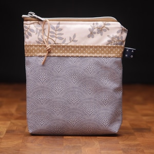 Könnte beinhalten: Ein rechteckiger Stoffbeutel mit Reißverschluss. Der Beutel hat oben ein hellgraues und weißes Blumenmuster, ein gepunktetes Band in Beige und unten ein graues fächerförmiges Muster. Ein kleines Etikett mit Text ist angebracht.