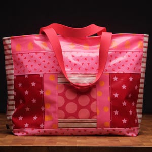 Könnte beinhalten: Eine rosa Tragetasche mit einem Patchwork-Design. Die Tasche weist verschiedene Muster auf, darunter Herzen, Sterne und Tupfen, in Rosa- und Rottönen. Sie hat einen einfarbigen rosa Griff und ist vor einem dunklen Hintergrund platziert.