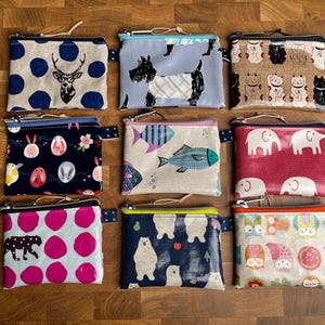 Könnte beinhalten: Neun kleine Reißverschlusstaschen mit verschiedenen Mustern werden auf einer Holzoberfläche präsentiert. Zu den Designs gehören Hirsche, Hunde, Katzen, Fische, Elefanten, Ostereier, Eisbären, ein Leopard und Eulen. Jede Tasche hat einen farbigen Reißverschluss.