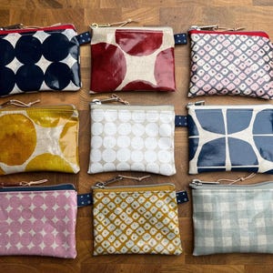 Könnte beinhalten: Neun rechteckige Reißverschlusstaschen in einem Raster angeordnet. Jede Tasche hat ein einzigartiges Muster, darunter große marineblaue Kreise, rote Kreise, rosa geometrische Formen, gelbe Kreise, weiße Punkte, blaue Kreuzformen, rosa Punkte, ein gelbes Gitter und hellblaues Vichy-Karo.