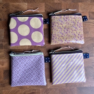 Könnte beinhalten: Vier kleine, quadratische Münzbörsen mit Reißverschlüssen und Stoffschlaufen. Die Geldbörsen weisen verschiedene Muster auf: große lila Punkte, kleine lila Blumen, winzige lila Sternchen und lila und weiße diagonale Streifen. Jede hat ein kleines blaues Etikett.