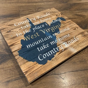 Pu&ograve; includere: Segno di legno con una silhouette blu dello stato della Virginia Occidentale. Il segno ha le parole "Country Roads take me home to the place I belong West Virginia mountain momma take me home Country Roads" scritte in bianco.