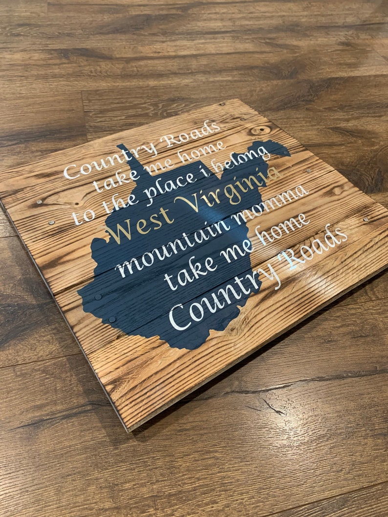 Pu&ograve; includere: Segno di legno con una silhouette blu dello stato della Virginia Occidentale. Il segno ha le parole "Country Roads take me home to the place I belong West Virginia mountain momma take me home Country Roads" scritte in bianco.