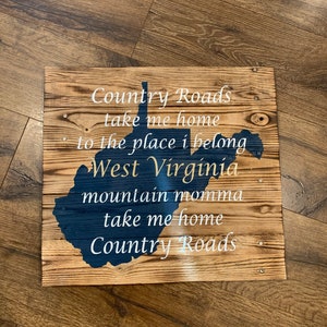 Pu&ograve; includere: Un cartello di legno con il contorno della Virginia Occidentale in vernice blu. Il cartello ha la scritta "Country Roads take me home to the place I belong West Virginia mountain momma take me home Country Roads" in vernice bianca.