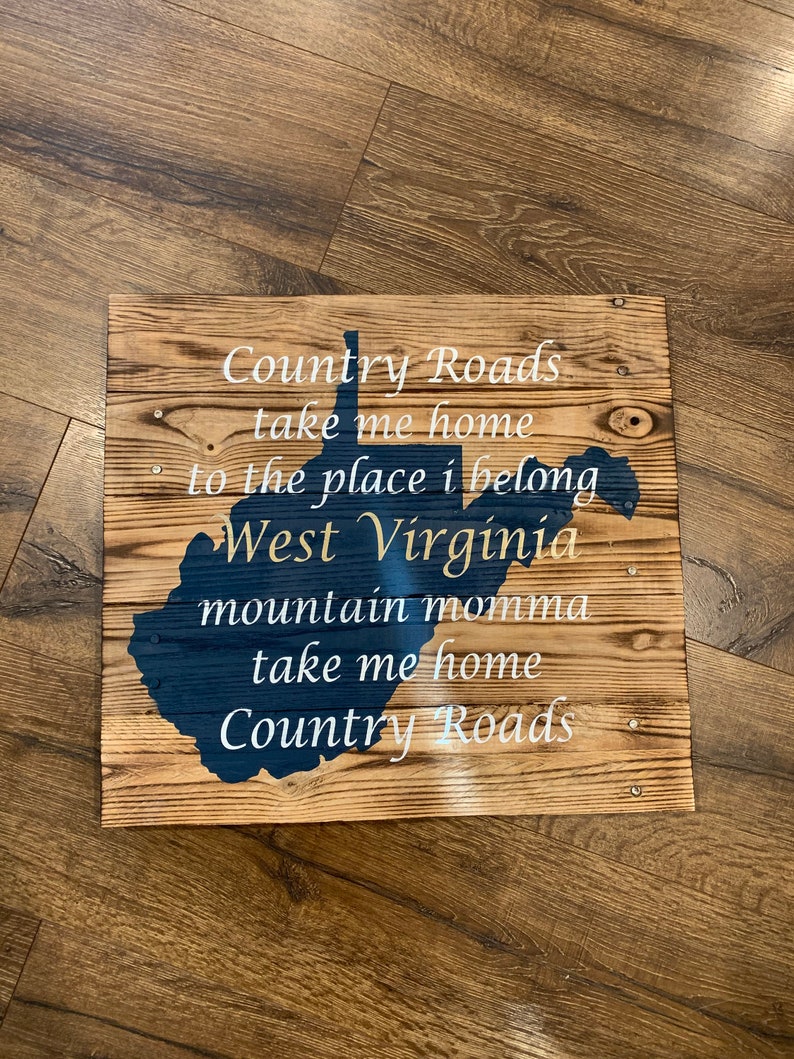 Pu&ograve; includere: Un cartello di legno con il contorno della Virginia Occidentale in vernice blu. Il cartello ha la scritta "Country Roads take me home to the place I belong West Virginia mountain momma take me home Country Roads" in vernice bianca.