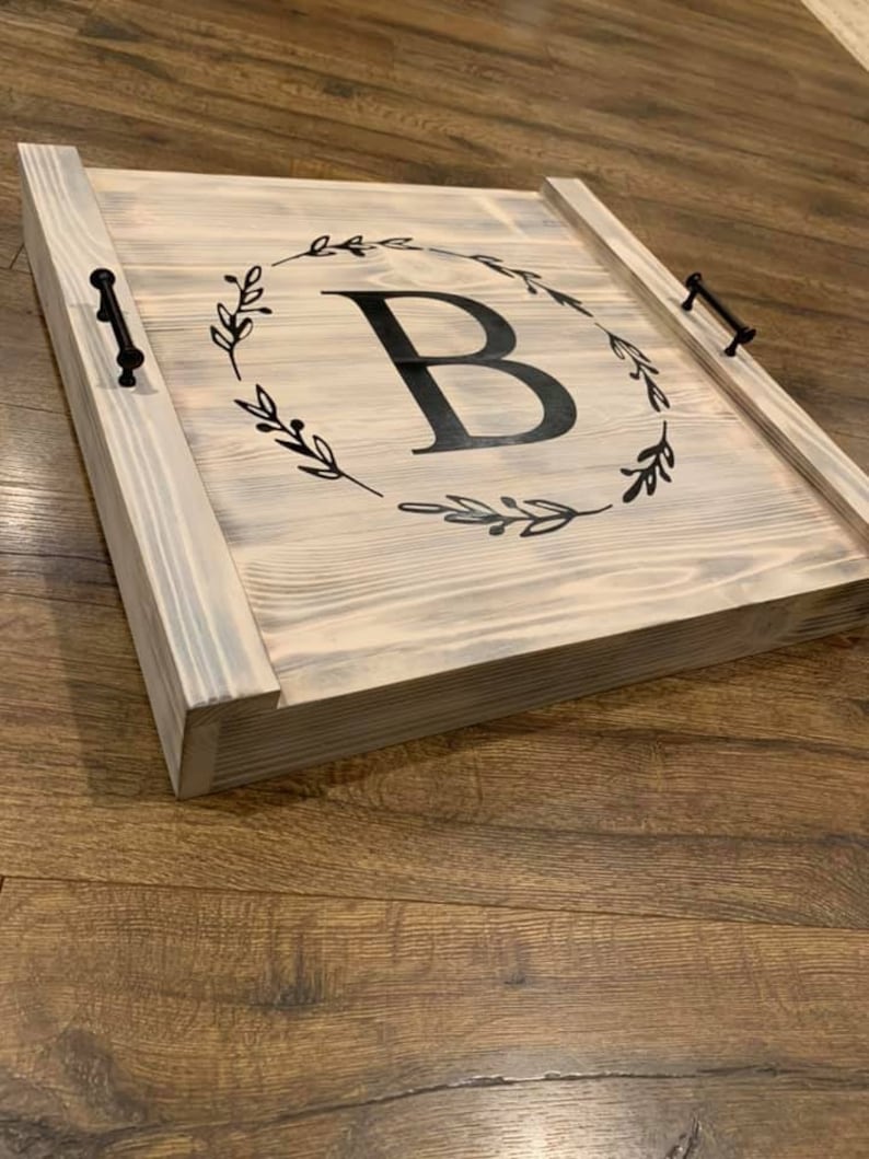 K&ouml;nnte beinhalten: Ein Holztablett mit grauem Anstrich und schwarzen Metallgriffen. Das Tablett zeigt ein schwarz-wei&szlig;es Design eines Kranzes mit Bl&auml;ttern und dem Buchstaben "B" in der Mitte.