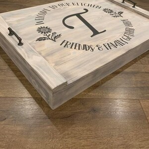 K&ouml;nnte beinhalten: Ein Holztablett mit wei&szlig; get&uuml;nchter Oberfl&auml;che. Das Tablett hat zwei schwarze Metallgriffe und eine schwarze Vinyl-Inschrift mit dem Text "Willkommen in unserer K&uuml;che, Freunde & Familie treffen sich hier". Die Inschrift befindet sich in einem Kreis mit einem gro&szlig;en Buchstaben "T" in der Mitte.