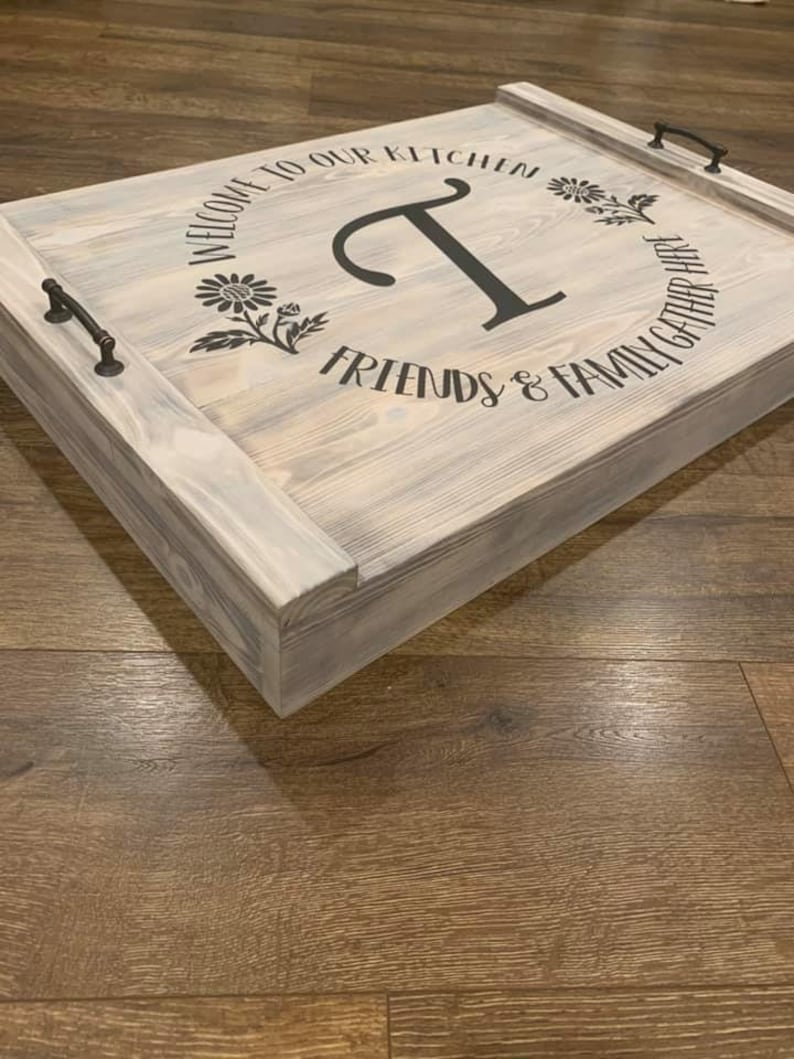 K&ouml;nnte beinhalten: Ein Holztablett mit wei&szlig; get&uuml;nchter Oberfl&auml;che. Das Tablett hat zwei schwarze Metallgriffe und eine schwarze Vinyl-Inschrift mit dem Text "Willkommen in unserer K&uuml;che, Freunde & Familie treffen sich hier". Die Inschrift befindet sich in einem Kreis mit einem gro&szlig;en Buchstaben "T" in der Mitte.