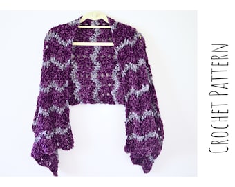 Può includere: Uno scialle all'uncinetto viola e grigio con un motivo geometrico. Lo scialle è appeso a un appendiabiti di legno su uno sfondo bianco. CROCHET PATTERN è scritto in testo nero sul lato destro dell'immagine.