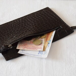 May include: A brown leather wallet with a zipper closure. The wallet is open and contains several bills and coins. The wallet has a crocodile-embossed pattern.