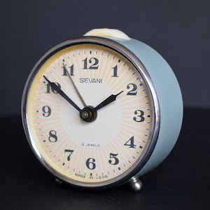 Pode incluir: Um despertador azul claro com um mostrador branco e ponteiros pretos. O despertador tem uma moldura prateada e diz "SEVANI" e "4 JEWELS" no mostrador. O despertador também diz "MADE IN U.S.A."