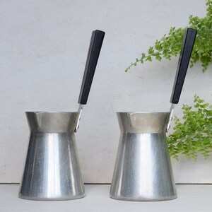 May include: Two silver coffee pots with black handles. The pots are on a white surface.