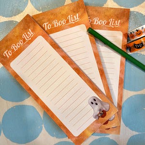 May include: Three Halloween-themed notepads with the text "To Boo List" in a vintage orange and white design. Each notepad has lined paper and a small ghost illustration. A green pen and decorative tape are also visible.
