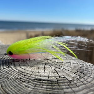 May include: A colorful fishing lure with a neon green body, pink accents, and white and green feathery extensions. The lure is set against a weathered wooden surface with a blurred ocean and sky background.