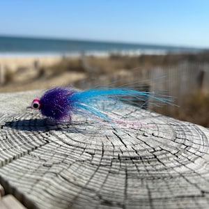 May include: A blue and purple fly fishing lure with pink accents. The lure is on a wooden surface with a blurred background of a beach and ocean.