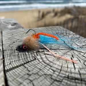 May include: A colorful fishing fly with a curved hook. The fly features a tan body, orange and blue accents, and a red eye. It is resting on a weathered wooden surface, with the ocean in the background.
