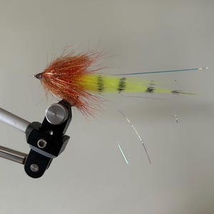 May include: A handmade fishing fly with a vibrant design. The fly features a reddish-brown head, transitioning to a yellow body with black stripes. Fine, iridescent strands extend from the fly, suggesting movement. It is mounted on a silver and black fly-tying vise.