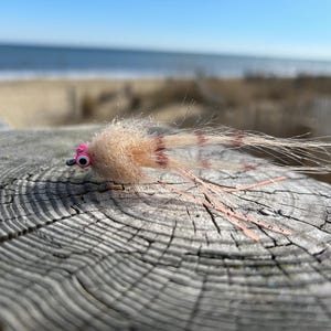 May include: A close-up of a light brown and pink fly fishing lure with a white and brown feather tail. The lure is sitting on a piece of weathered wood.