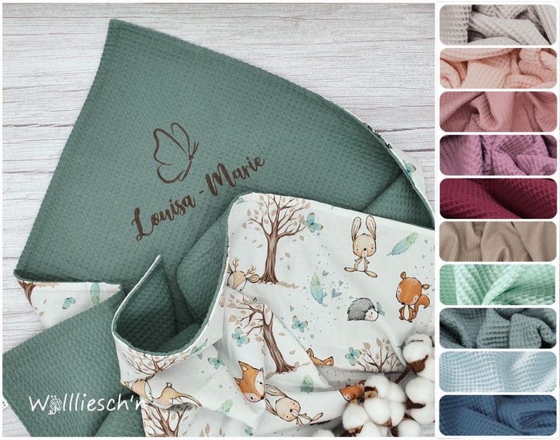 May include: A sage green waffle-knit blanket featuring a butterfly icon and the name Louisa-Marie embroidered in brown. The reverse side displays a white fabric with a pattern of trees, rabbits, hedgehogs, foxes, and feathers in soft colors.