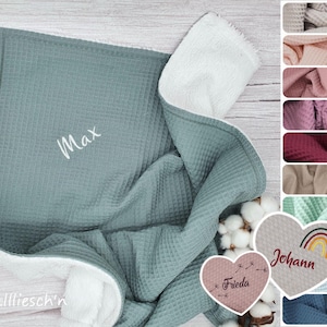 Könnte beinhalten: Eine weiche, grüne Waffel-Babydecke mit einem weißen Fleece-Rücken. Die Decke ist mit dem Namen "Max" in weißer Stickerei personalisiert. Die Decke ist in der Hälfte gefaltet und der weiße Fleece-Rücken ist sichtbar. Die Decke ist von weißen, flauschigen Blumen und einem herzförmigen Etikett mit dem Namen "Frieda" umgeben.