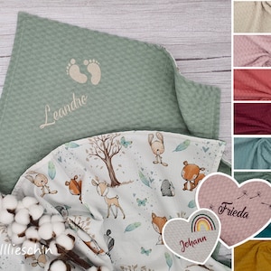 Könnte beinhalten: Zwei personalisierte Babydecken. Eine ist grün mit einem Fußabdruck-Design und dem Namen "Leandro" bestickt. Die andere ist weiß mit einem Waldtier-Druck und den Namen "Johann" und "Frieda" bestickt.