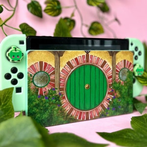 May include: A green and white Nintendo Switch console with a custom design featuring a green door with a red brick frame and a leafy green background. The design is inspired by The Lord of the Rings.