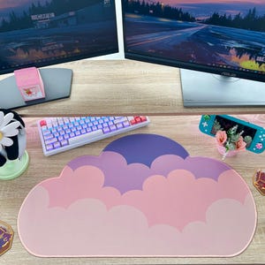 May include: A pink, purple, and white cloud-shaped mousepad sits on a wooden desk, next to a white keyboard with pink keys. A small pink strawberry milk carton and a daisy plushie are also visible.