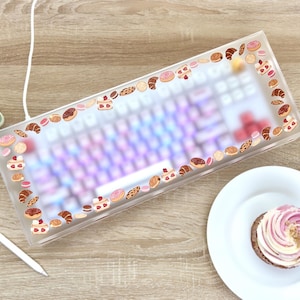 Peut inclure: Un clavier transparent en acrylique avec un clavier rose et blanc à l'intérieur. Le boîtier est décoré de petites images de pâtisseries et de desserts.