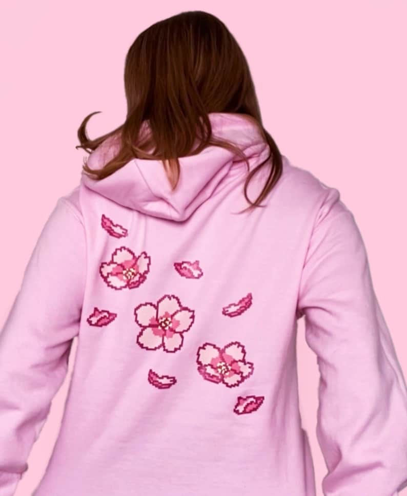Woman wearing a pink hoodie with screen printed pixel cherry blossoms and petals floating down the back over a pink background.