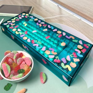May include: A teal blue keyboard case with a white keyboard inside. The case is decorated with colourful candy-like stickers. A bowl of colourful sweets is in the foreground.