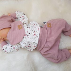 Könnte beinhalten: Eine Babypuppe trägt eine gerippte Mütze in Altrosa, ein passendes geripptes Lätzchen, ein langärmeliges Oberteil mit weißem Blumenmuster und eine gerippte Hose in Altrosa. Das Outfit hat kleine braune Etiketten mit einem Herzsymbol am Lätzchen.
