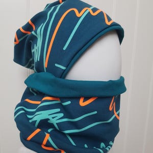 Op de afbeelding: Tealkleurige muts en nekwarmer set. De stof heeft een teal achtergrond met een abstract ontwerp in oranje en lichtblauw. De nekwarmer is omgeslagen en de muts is slouchy.