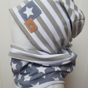 Könnte beinhalten: Ein Set aus grau-weiß gestreifter Beanie-Mütze und passendem Loop-Schal. Die Mütze hat einen grauen quadratischen Aufnäher mit einem weißen Stern und einen braunen Kunstlederaufnäher mit Sternaussparung. Der Schal hat einen grauen Hintergrund mit weißen Sternen.